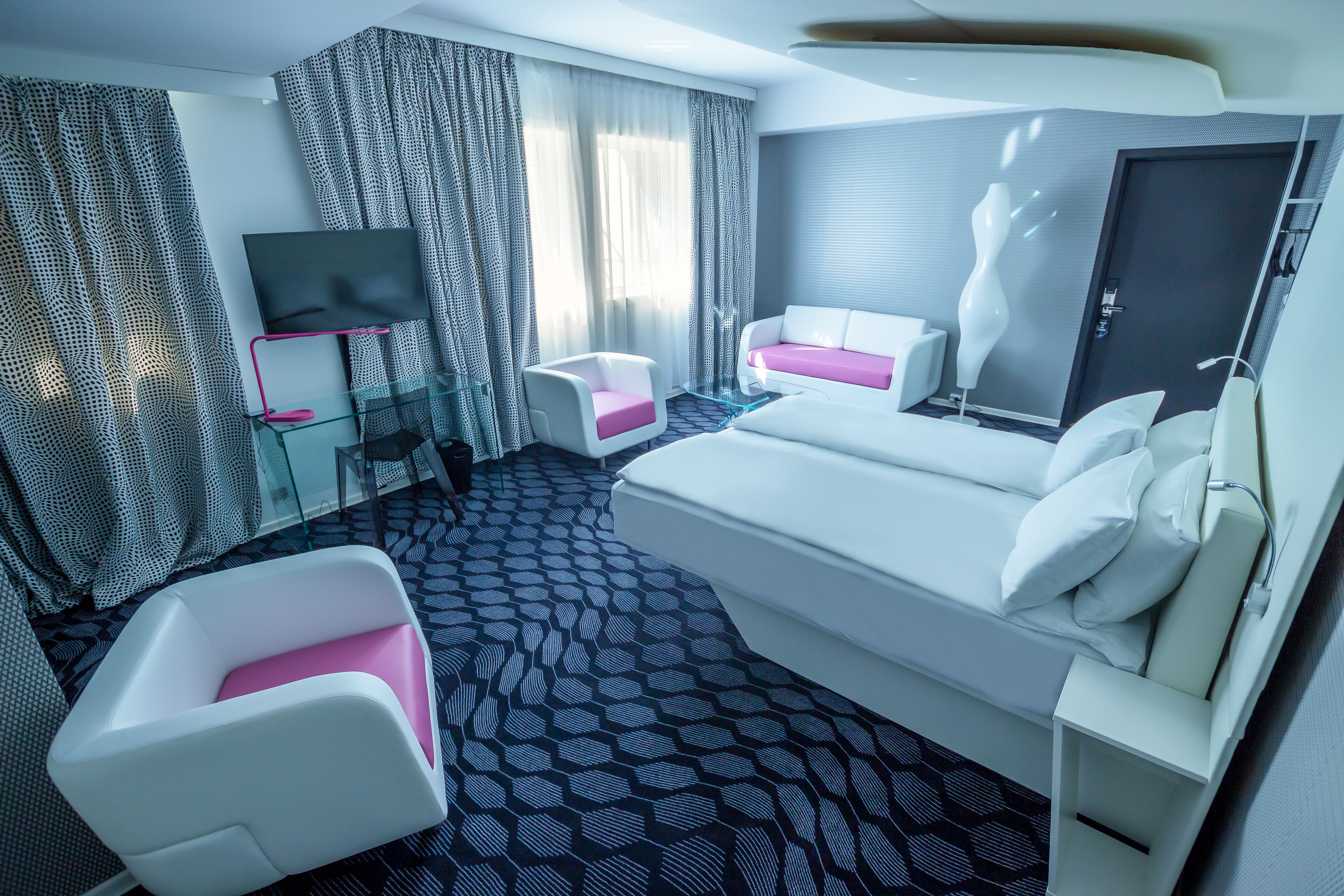 Een moderne hotelkamer met een groot bed, een zithoek met een roze en witte stoel, een flatscreen televisie en kamerbreed gordijn.