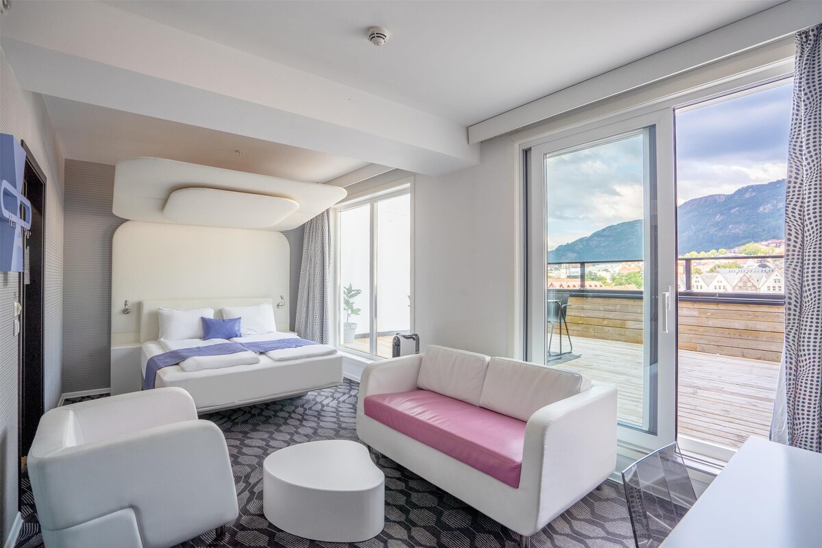 A modern hotel room with a bed, a sofa, a chair, and a glass table. There is a balcony with a view of the mountains.