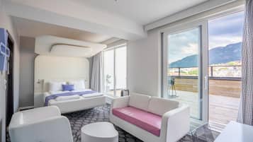 A modern hotel room with a bed, a sofa, a chair, and a glass table. There is a balcony with a view of the mountains.