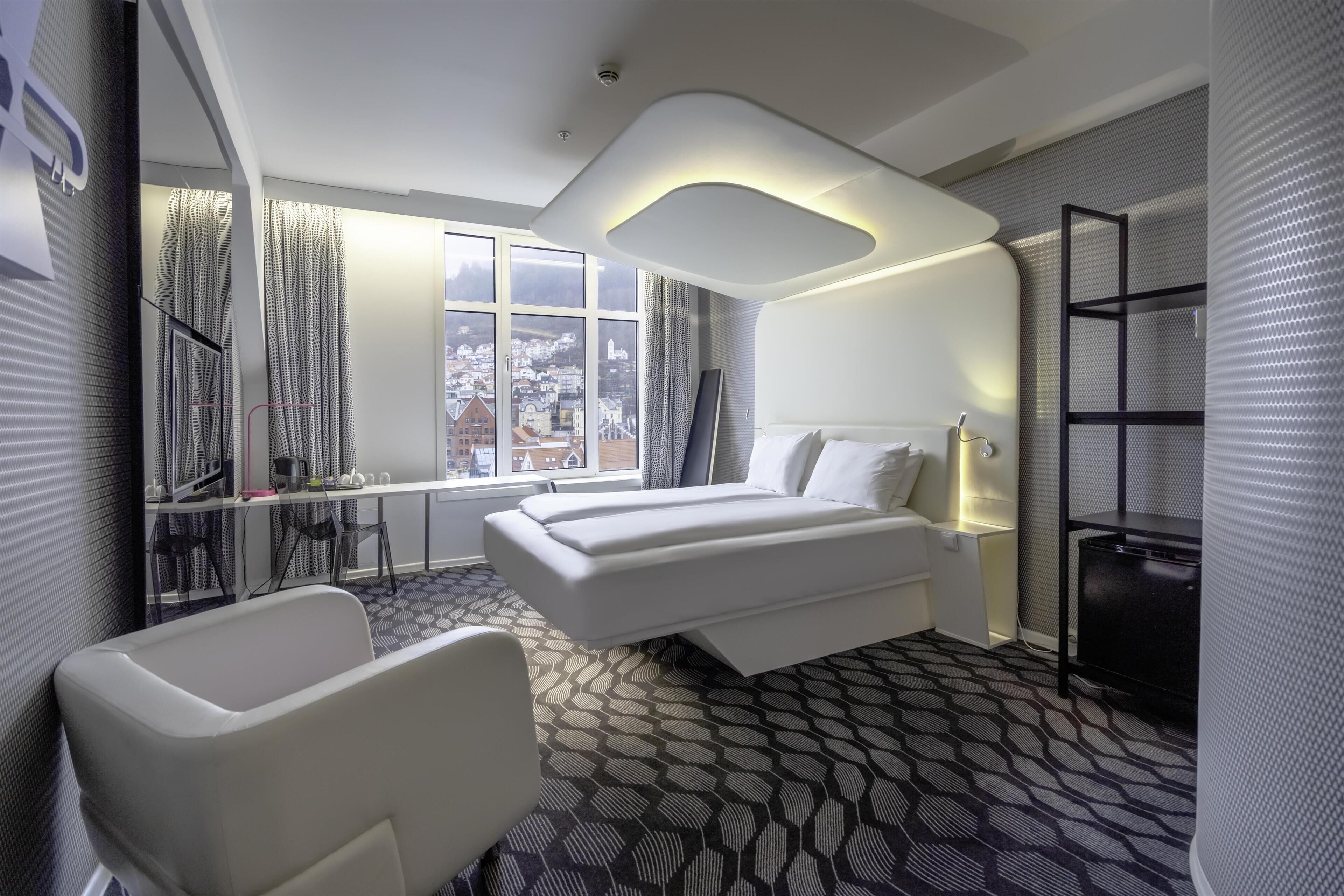 A modern hotel room with a large bed, a desk, a chair, and a view of the city.