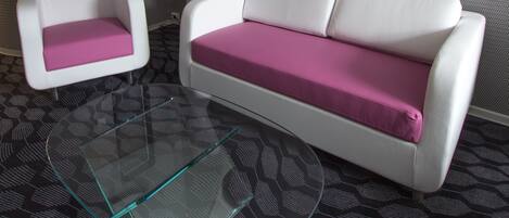A modern room with a white sofa and a white armchair, both with pink cushions, a glass coffee table, and a patterned carpet.