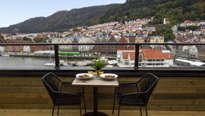 A balcony with a table and two chairs, overlooking a town with red-roofed buildings and a body of water.