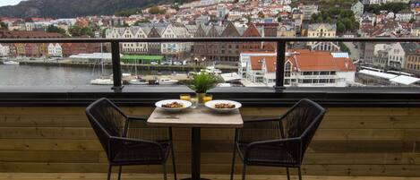 A balcony with a table and two chairs, overlooking a town with red-roofed buildings and a body of water.