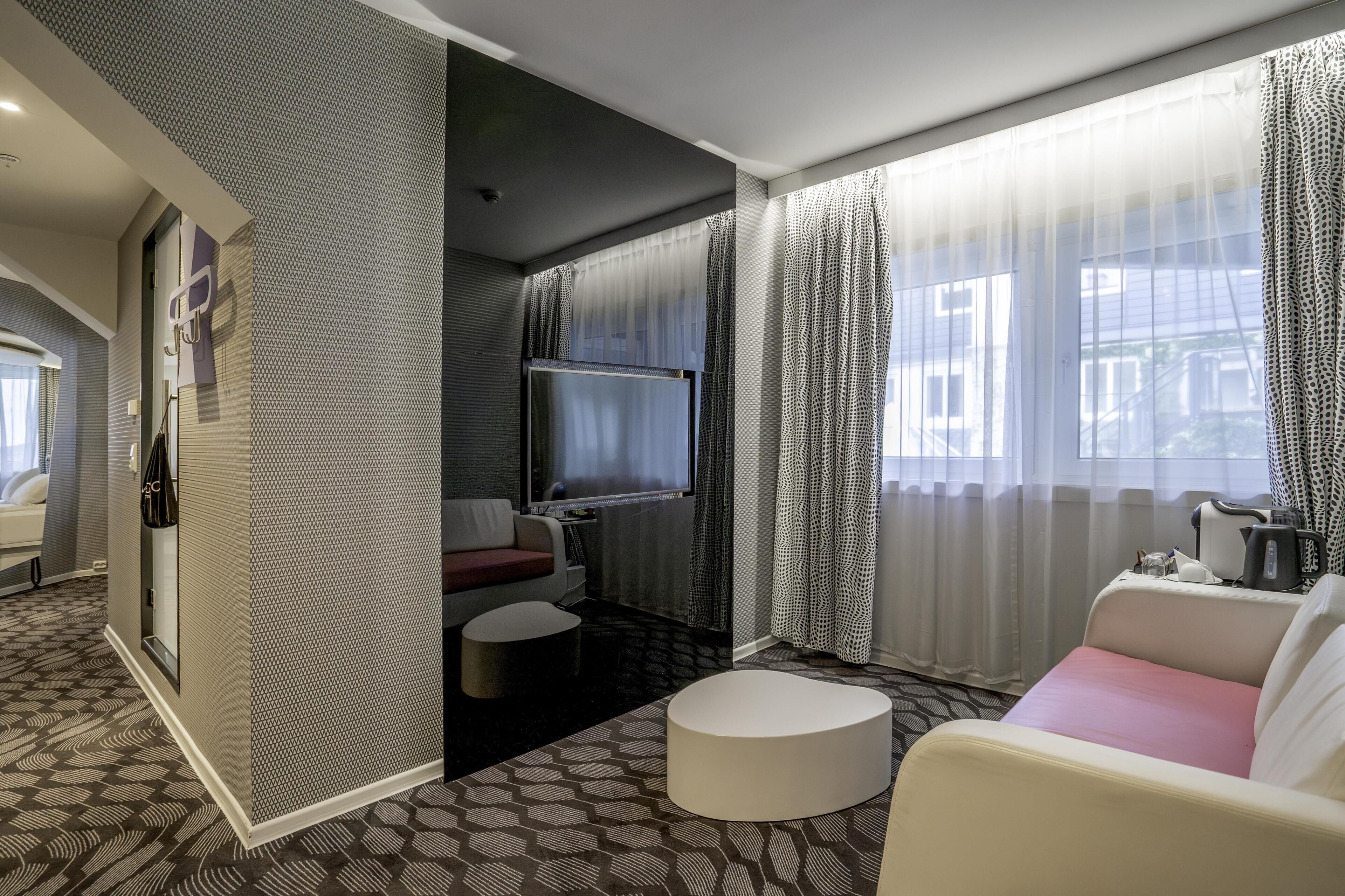 A hotel room with a sofa, a small round table, a TV, and a window with patterned curtains.