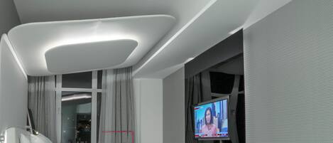 A modern living room with a glass coffee table, a white sofa with a pink cushion, and a flat-screen TV mounted on the wall.