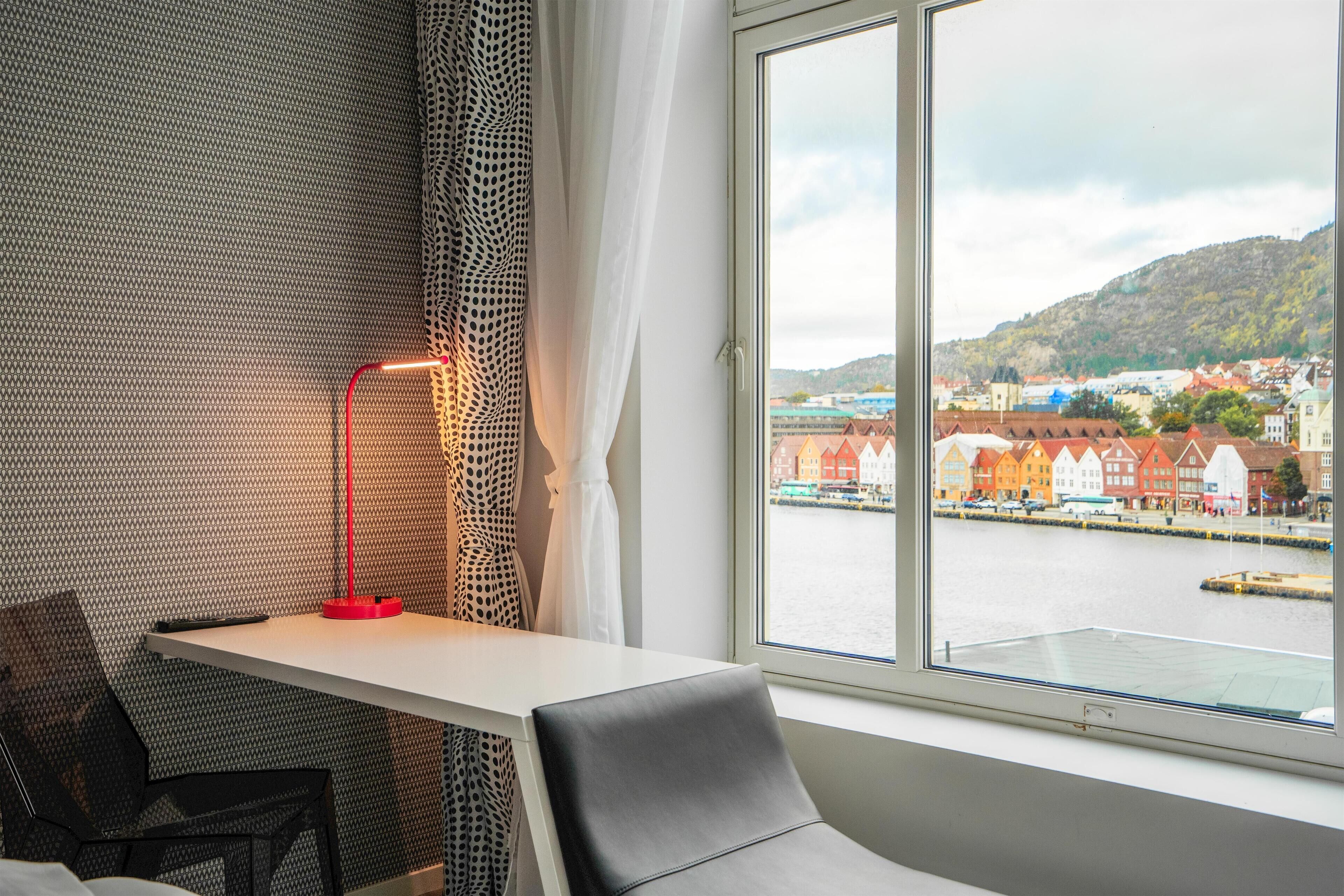 A room with a table, a chair, a red floor lamp, and a window with a view of a waterfront and buildings.