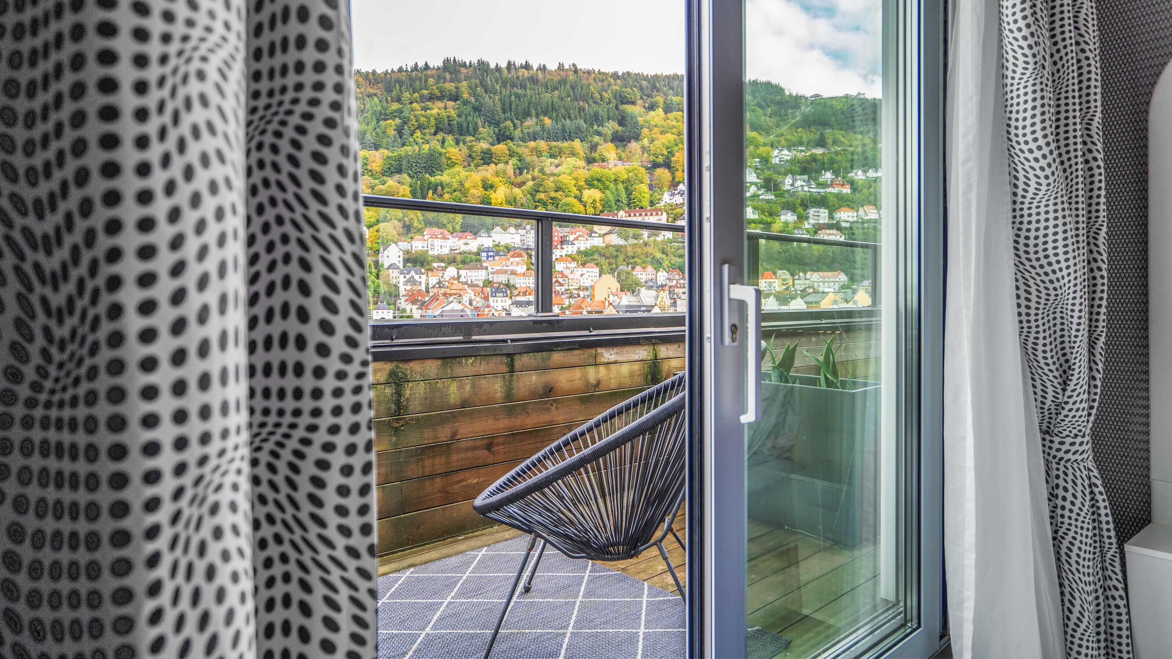 Een balkon met een rieten stoel, een glazen deur en uitzicht op een heuvelstad.