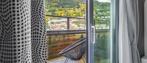 Een balkon met een rieten stoel, een glazen deur en uitzicht op een heuvelstad.