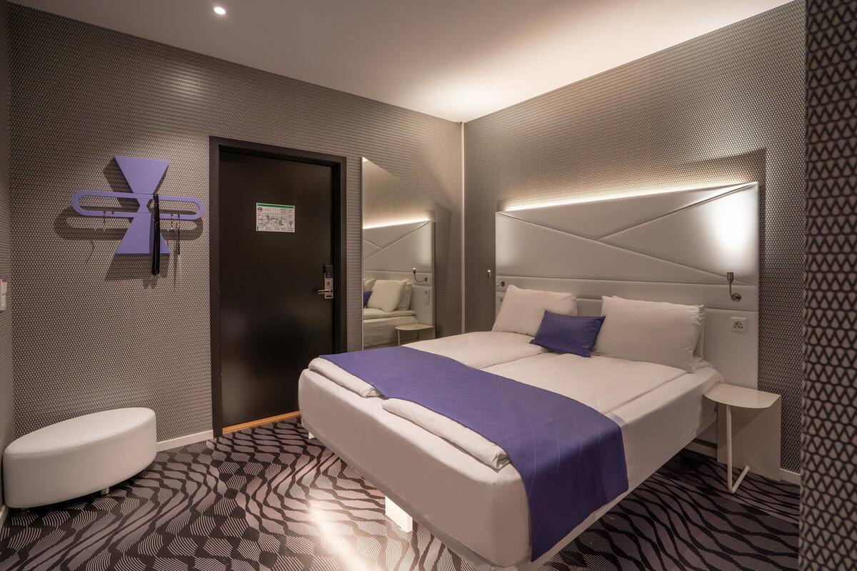 Une chambre d’hôtel moderne avec un grand lit, une table de chevet, un petit coin salon circulaire et un miroir au-dessus du lit.