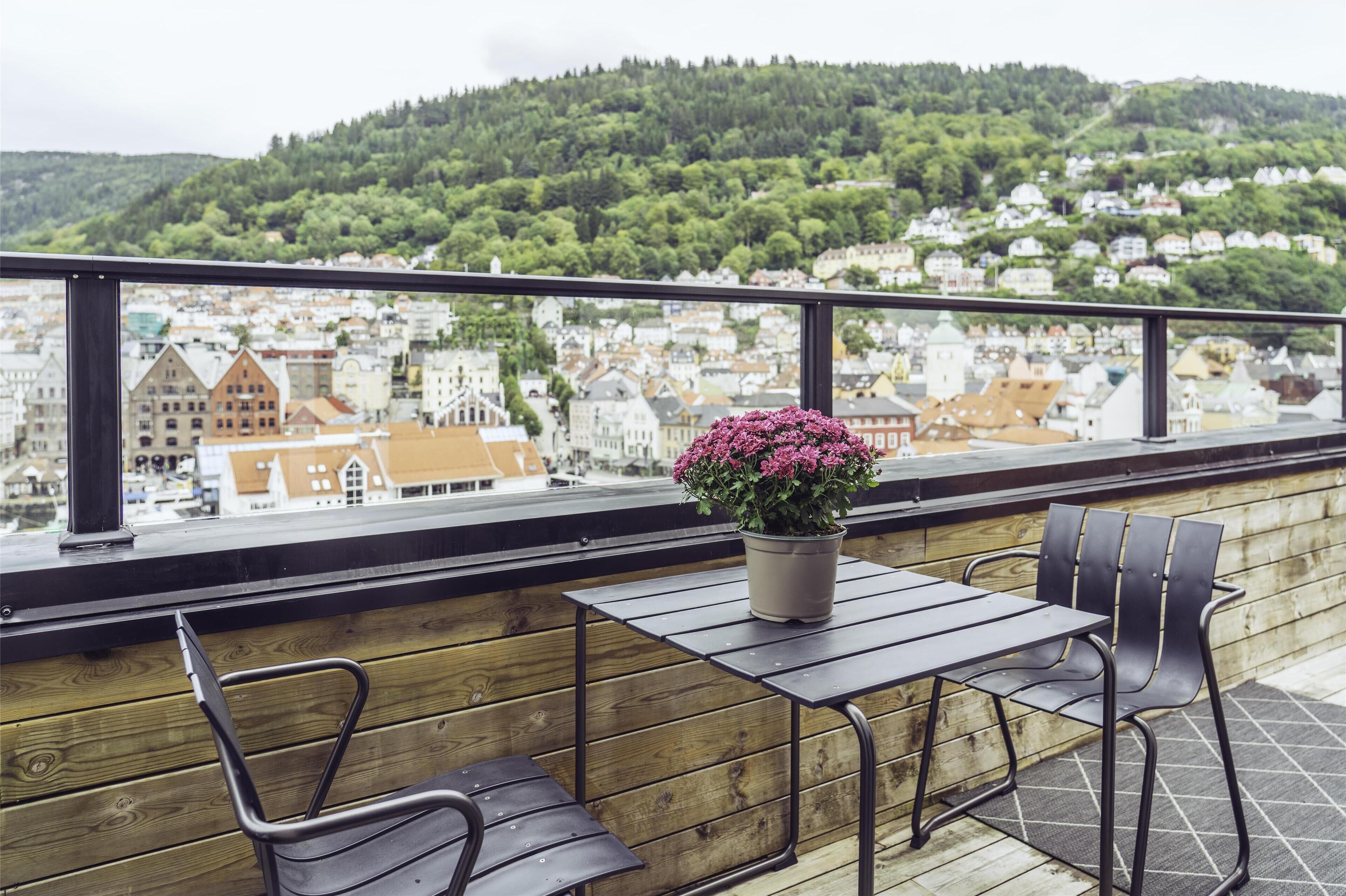A balcony with a wooden railing, a table, two chairs, and a potted plant with pink flowers.