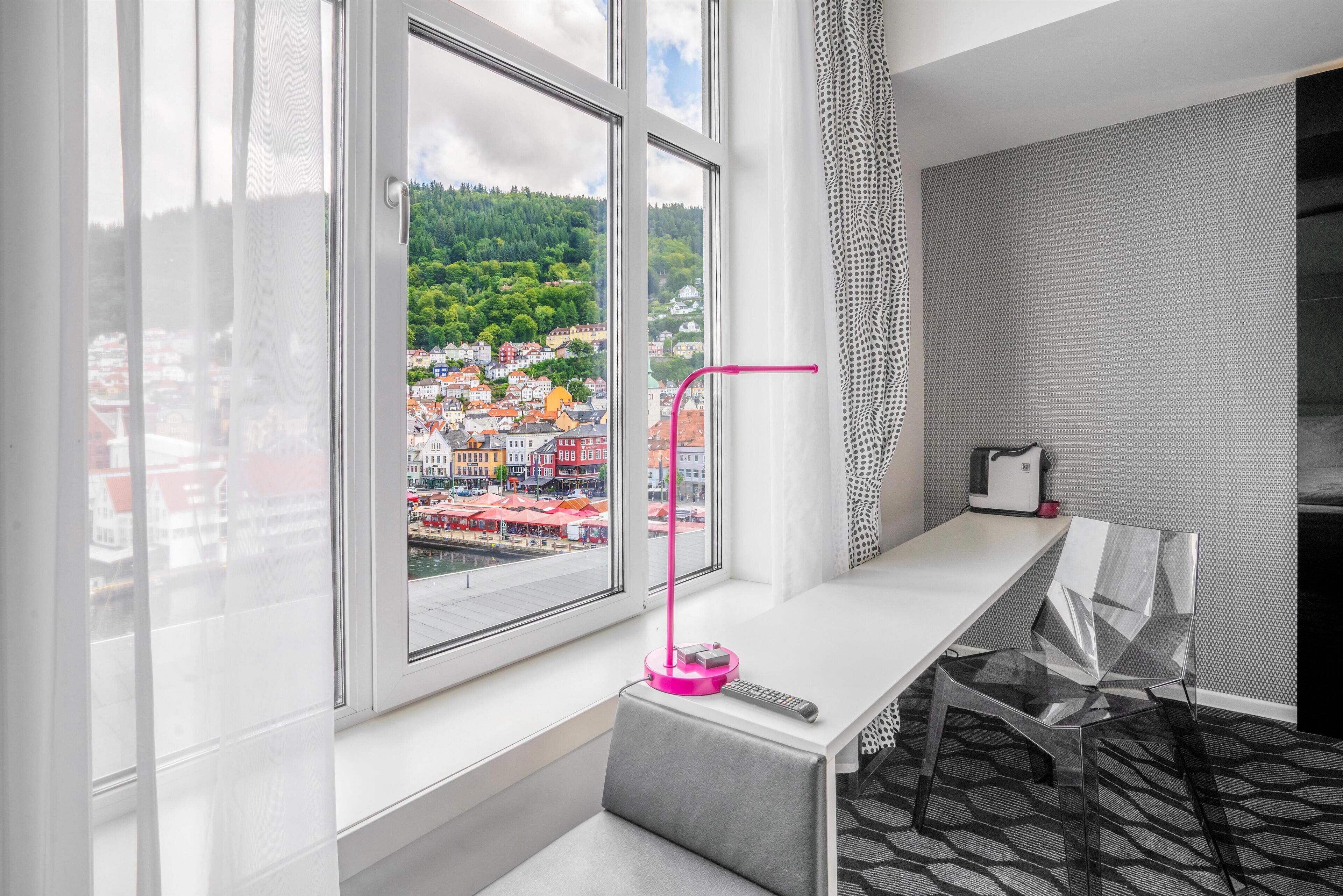 Habitación de hotel moderna con un amplio ventanal que ofrece vistas a un pueblo y colinas. Se observa un escritorio con una silla, una lámpara rosa y un control remoto en el alféizar.