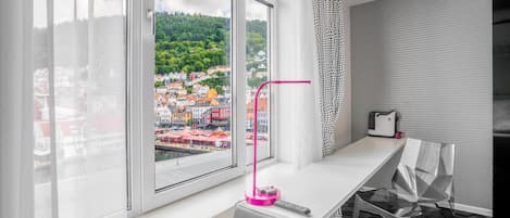 Habitación de hotel moderna con un amplio ventanal que ofrece vistas a un pueblo y colinas. Se observa un escritorio con una silla, una lámpara rosa y un control remoto en el alféizar.