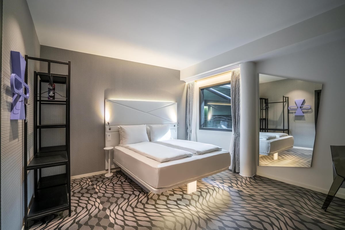 Une chambre d’hôtel moderne comprenant un lit, une tête de lit dotée d’un éclairage intégré, une étagère noire, une moquette à motifs et un grand miroir qui reflète la pièce.