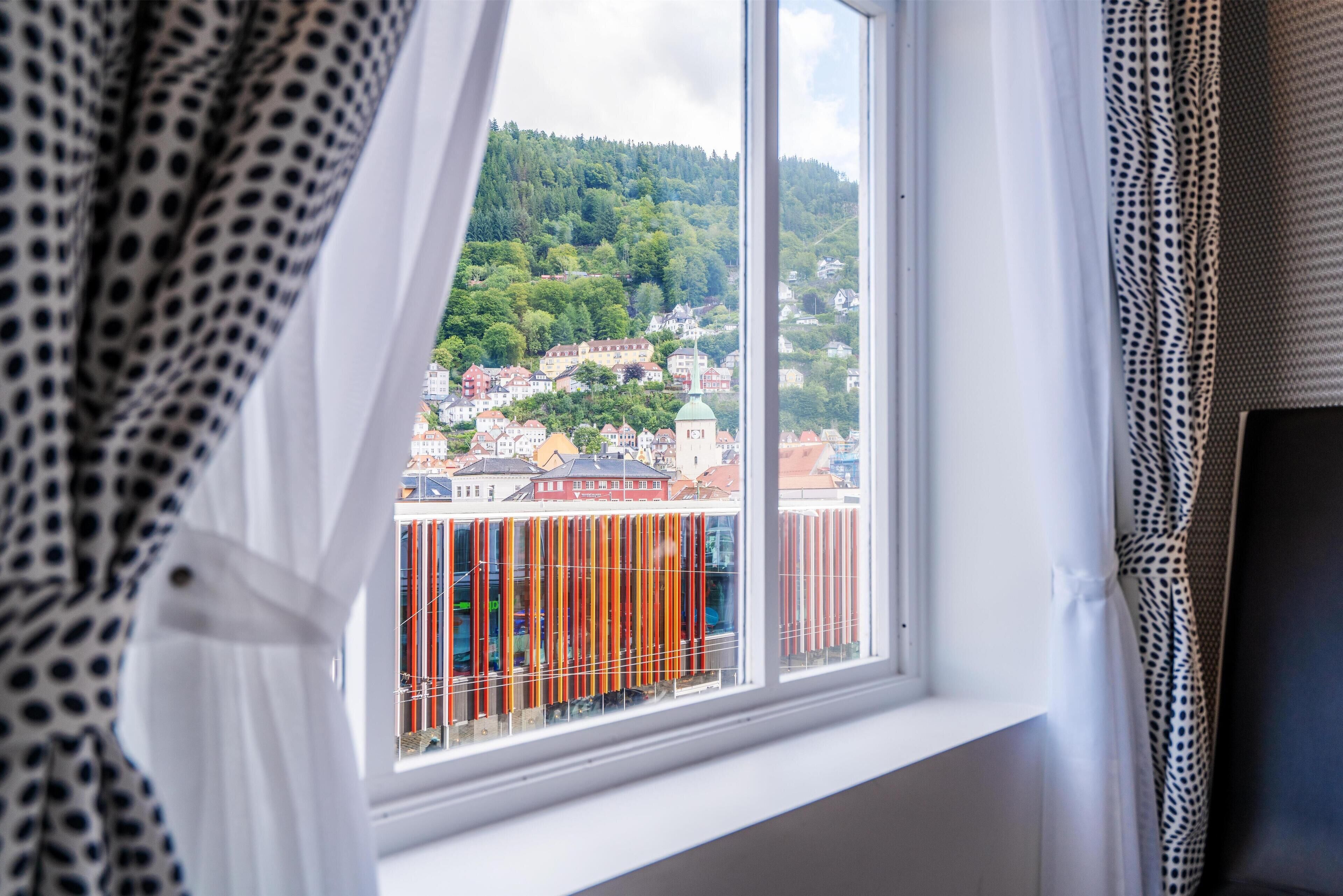 A window with a view of a town, including buildings and a green hill.
