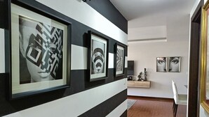 Un couloir aux murs rayés de noir et de blanc, orné de tableaux encadrés, doté d’un plancher en bois et comprenant un coin repas avec une chaise et une table.