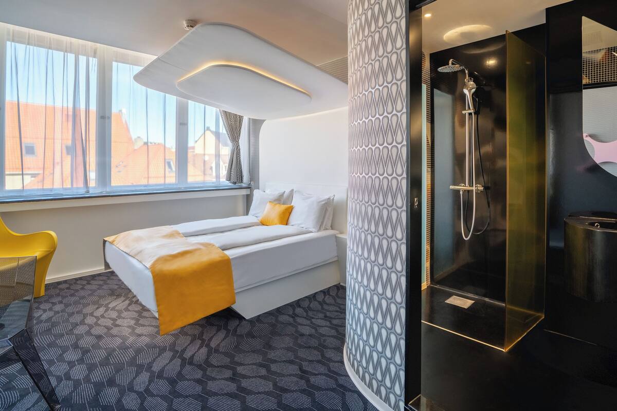 A modern hotel room with a bed, a shower, and a view of buildings outside.