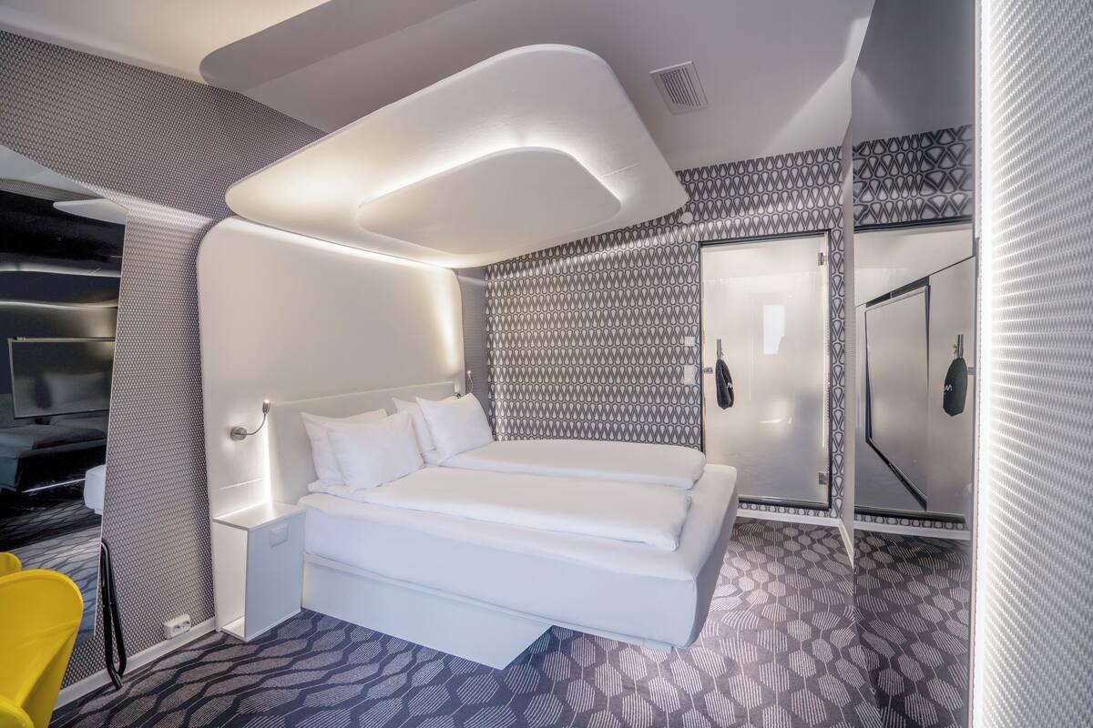 A modern hotel room with a large bed, a bedside table, a mirror, and a patterned floor.