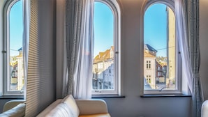 Una habitación con sofá, una mesa de centro de vidrio y tres ventanas amplias con cortinas.