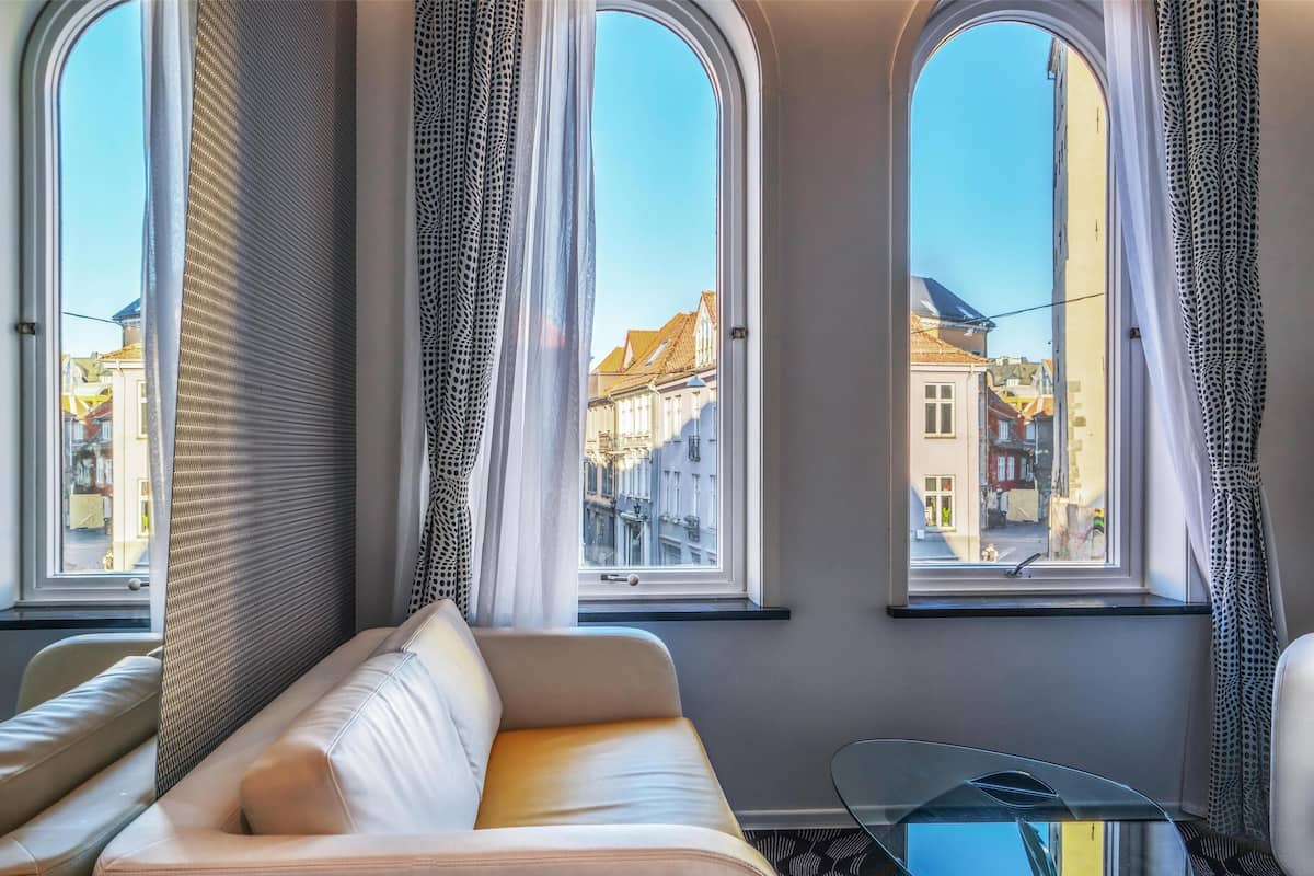 A room with a sofa, a glass coffee table, and three large windows with curtains.