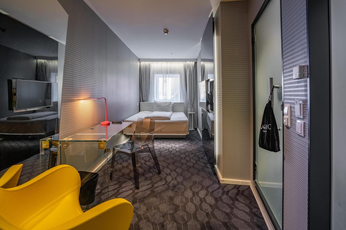 A modern hotel room with a bed, a desk with a chair, a TV, and a glass partition.