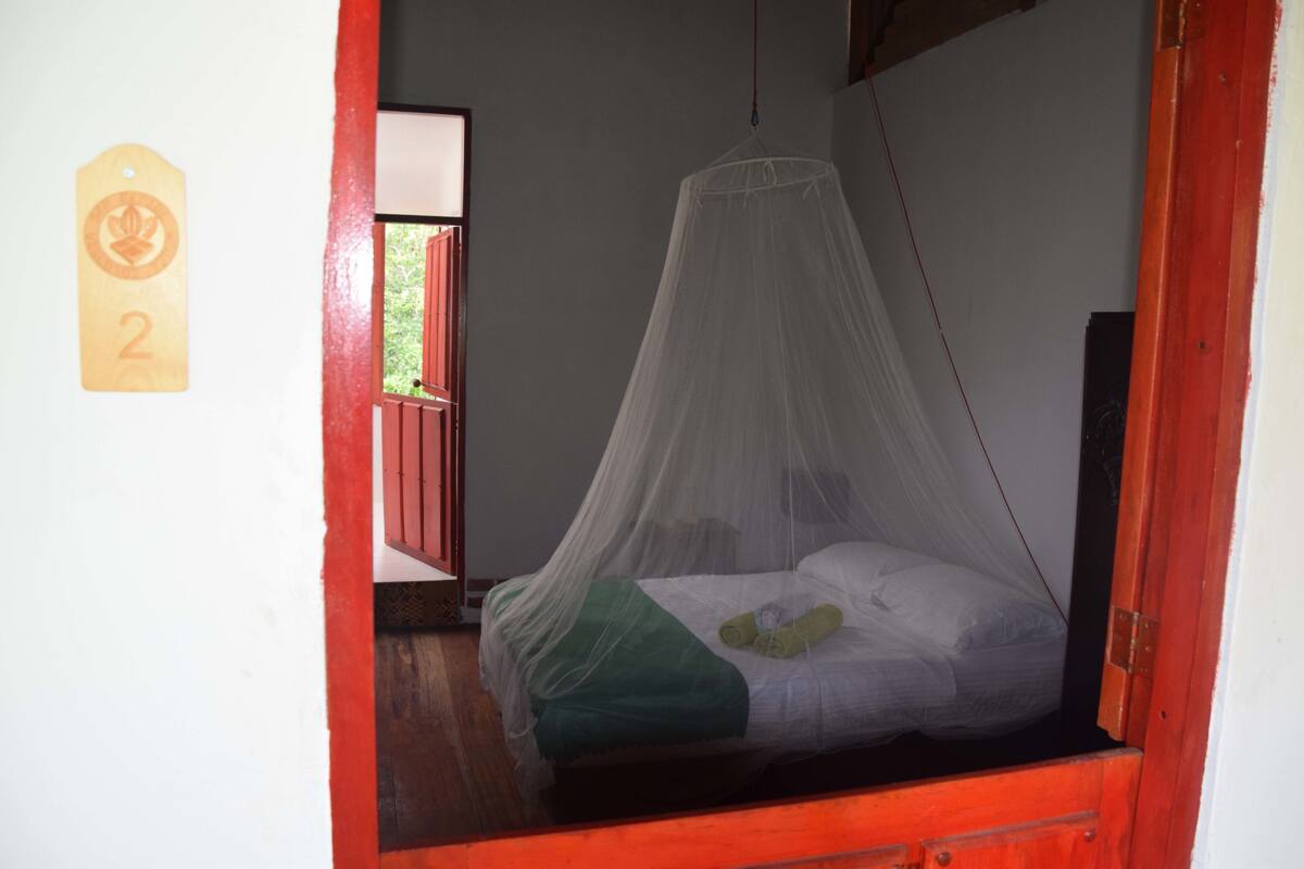 A room with a bed under a mosquito net, a wooden door, and a wall-mounted number tag.