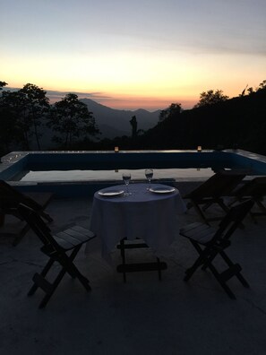 A sunset view from a poolside table with two chairs, a table set with plates and glasses, and a candle on the table. - Finca Cafetera San Rafael (Santa Marta)