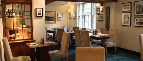 A dining area with wooden tables and chairs, framed pictures on the walls, and a wine display cabinet.