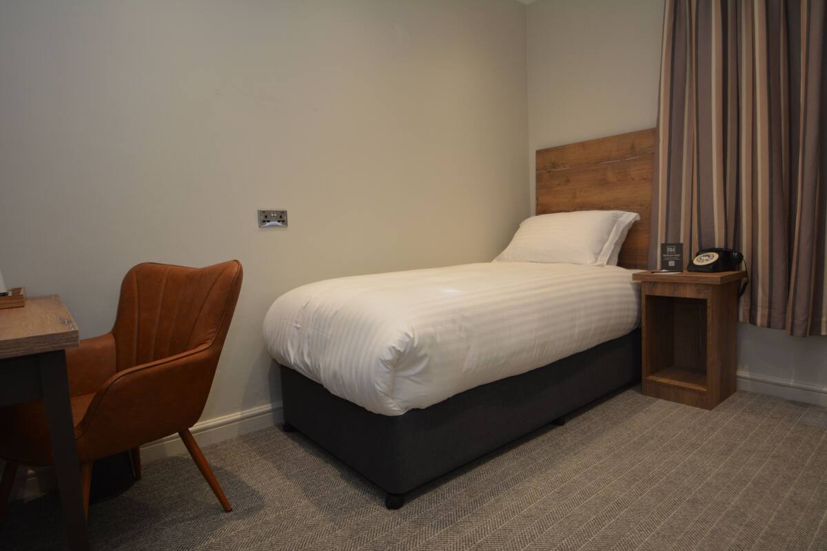 A hotel room with a single bed, a wooden nightstand, a chair, and a desk.