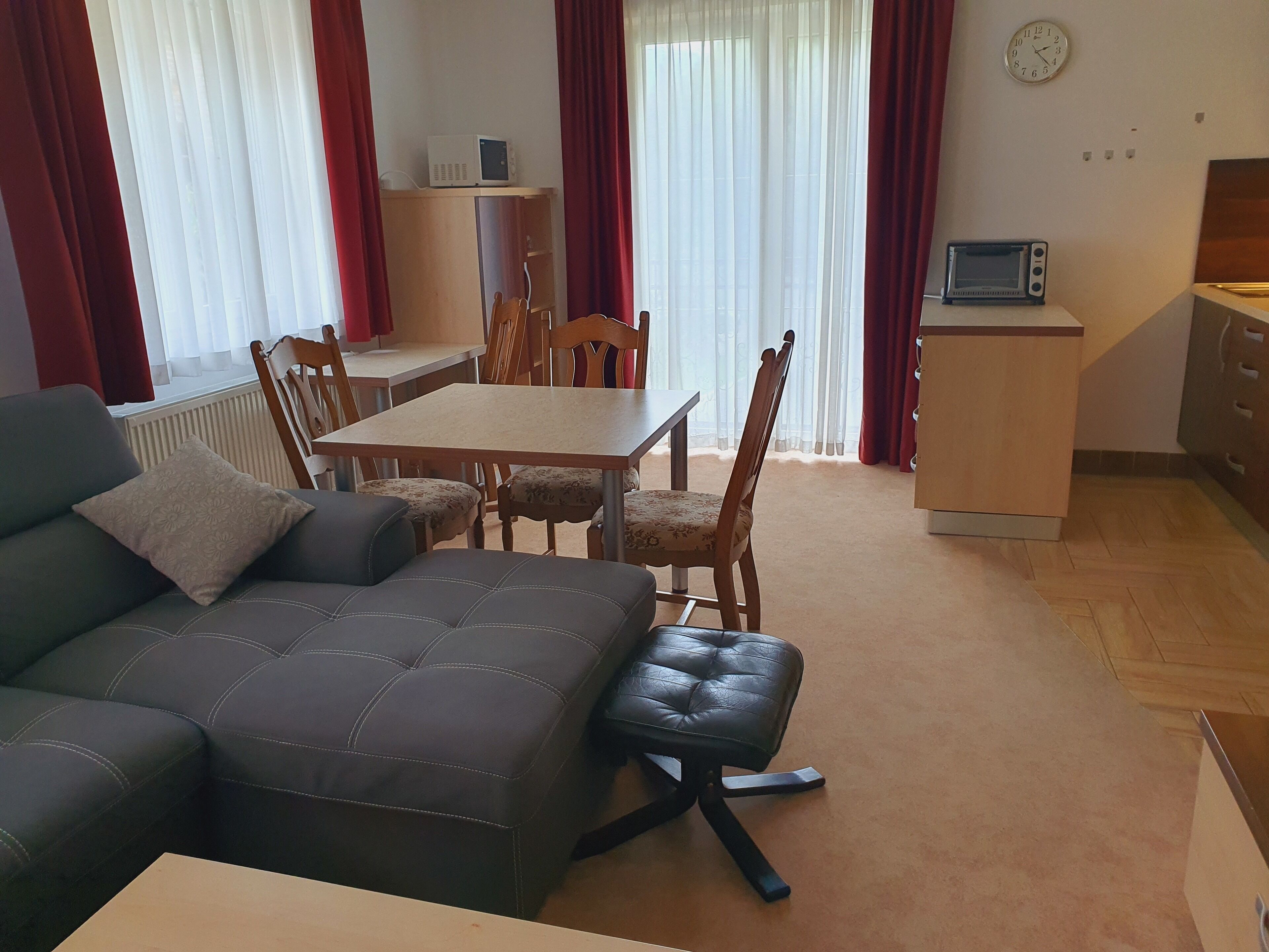 A living room with a sofa, a dining table with chairs, a small kitchen area with a microwave, and a clock on the wall.