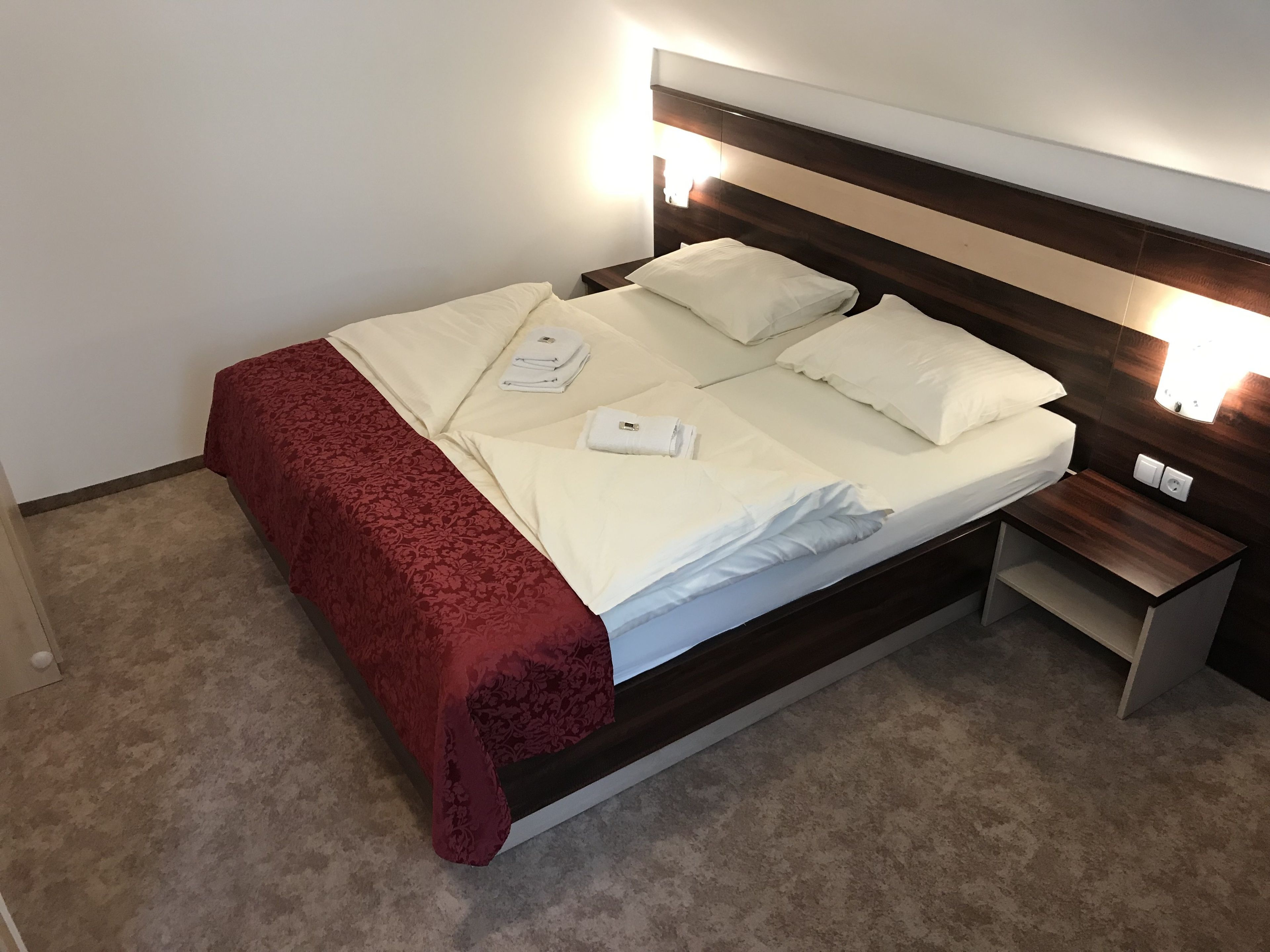 Un lit bien fait, avec du linge de lit blanc et un plaid rouge, une table de chevet avec une lampe, et un luminaire fixé au mur.