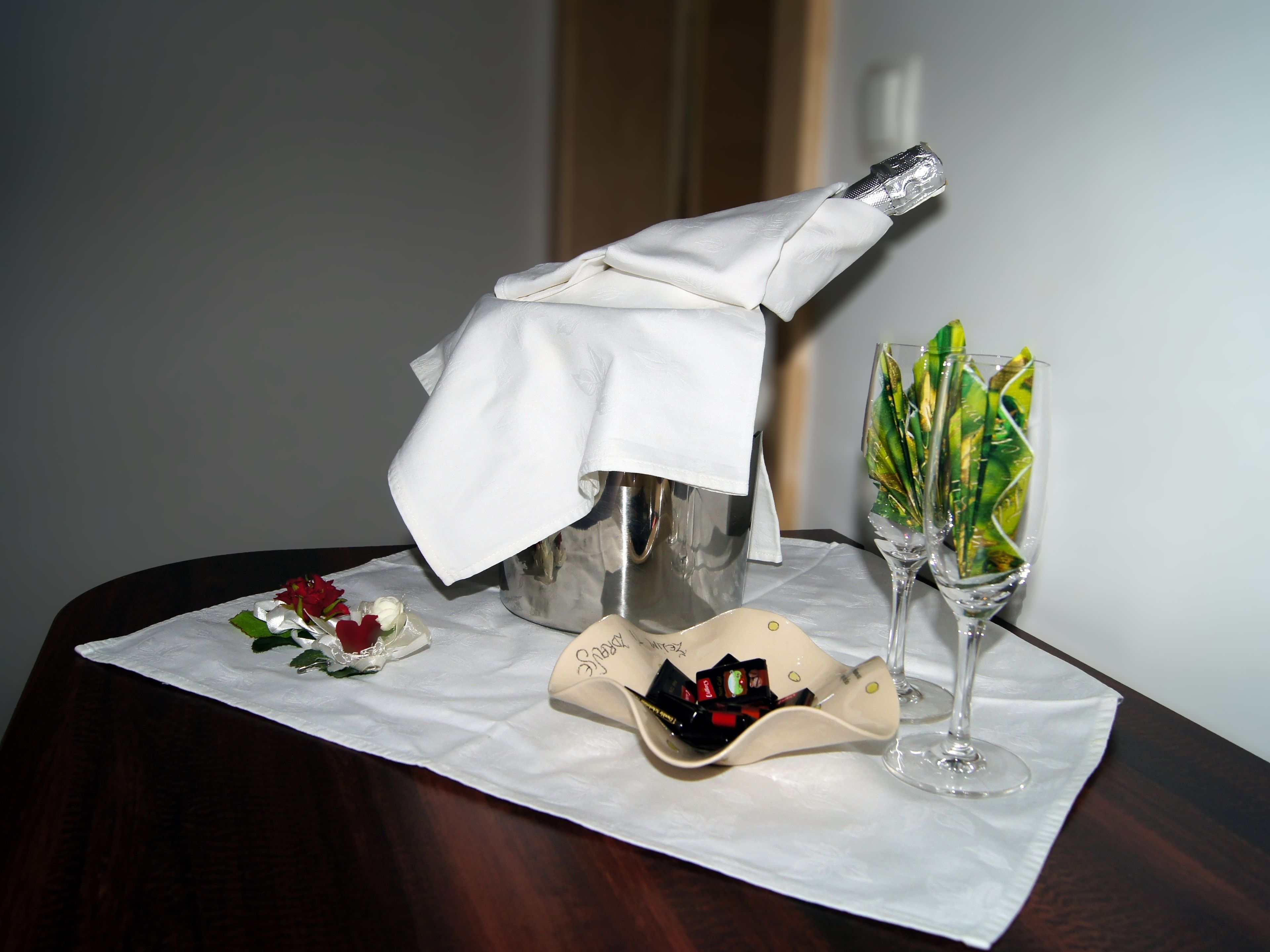 A table with a bottle of champagne, two glasses of wine, a small dish with chocolates, and a decorative flower arrangement.