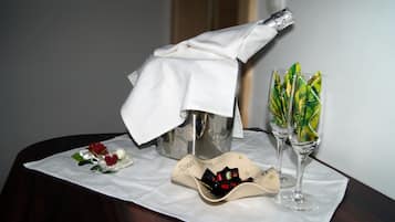 A table with a bottle of champagne, two glasses of wine, a small dish with chocolates, and a decorative flower arrangement.