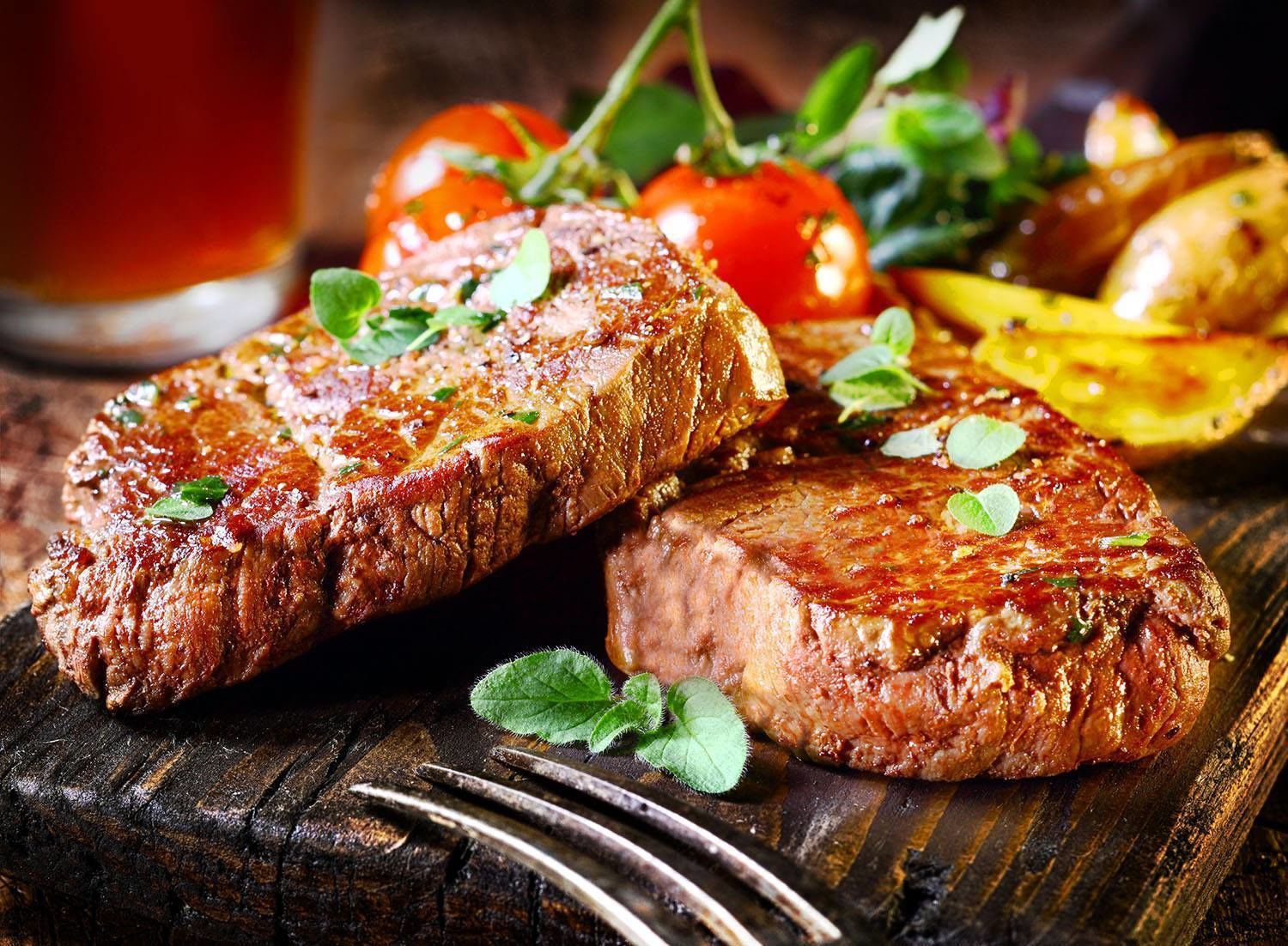 Twee gegrilde steaks, versierd met kruiden, geserveerd met geroosterde aardappelen en tomaten op een houten ondergrond.