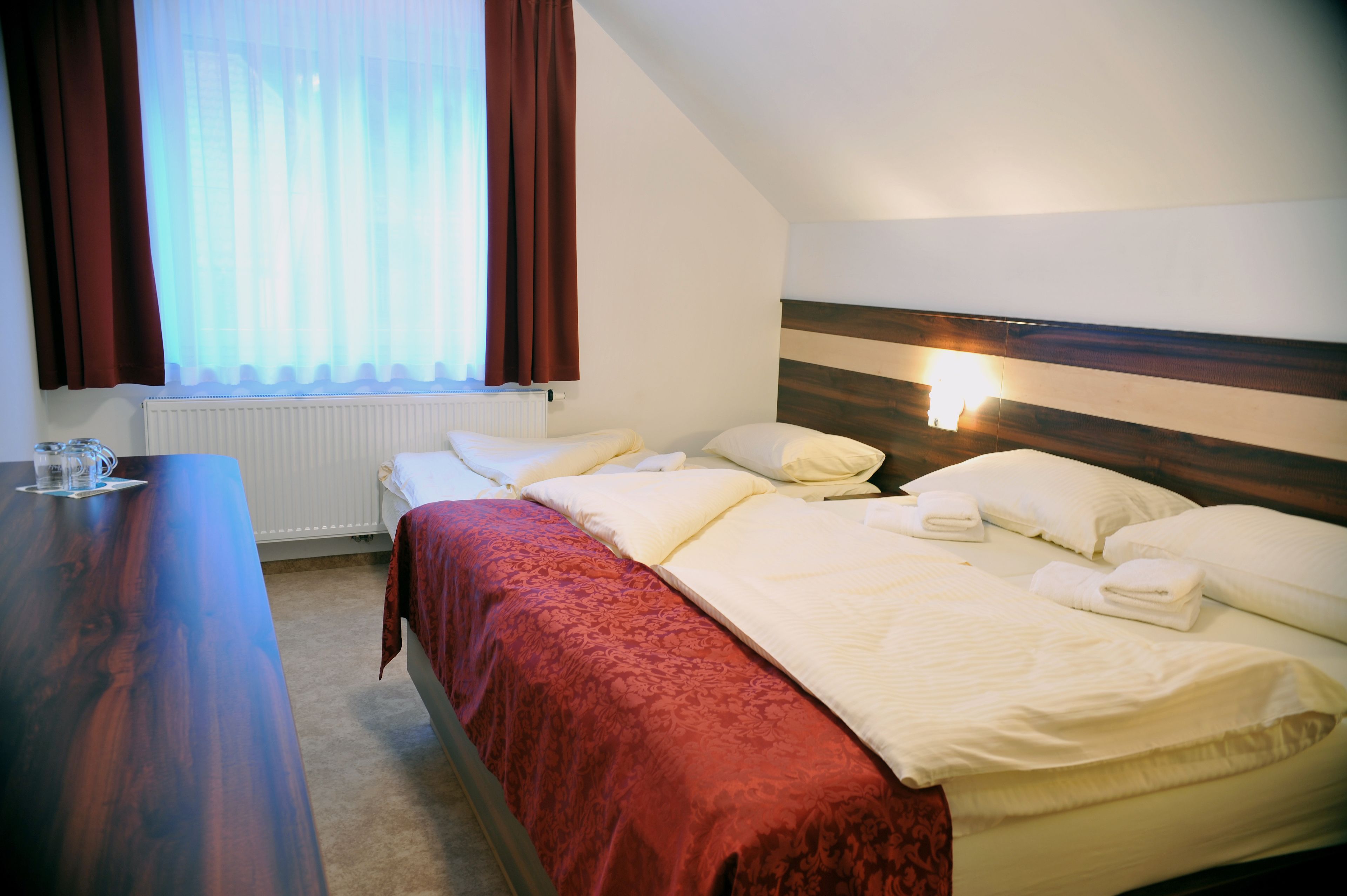 Quarto de hotel com escrivaninha de madeira, duas camas com roupa de cama vermelha e branca, radiador e janela com cortinas.