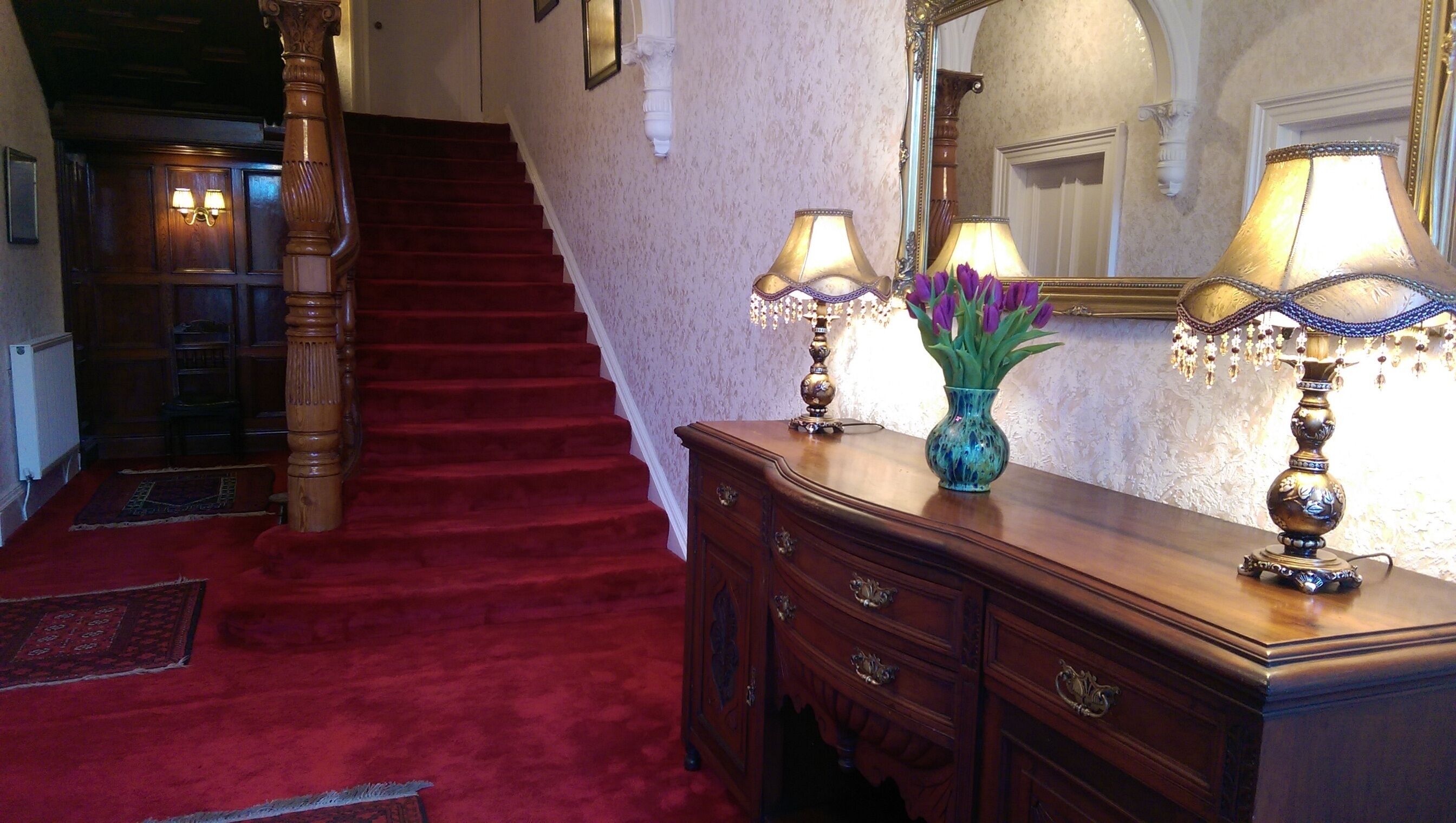 A grand staircase with red carpet, wooden balustrades, and wall-mounted lamps.