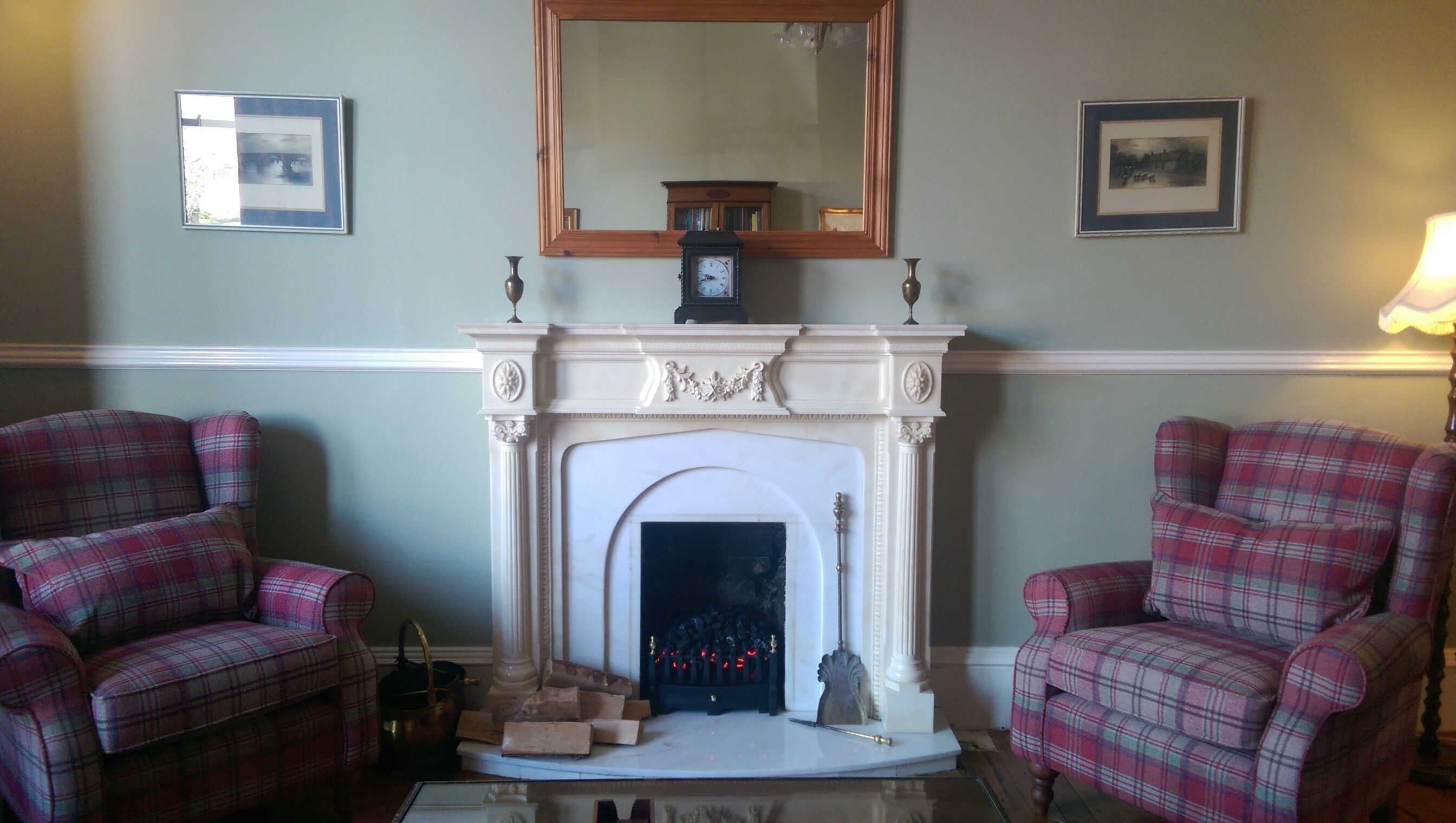 Una habitación con chimenea, dos sillones a cuadros, una mesa con lámpara y cuadros en las paredes.