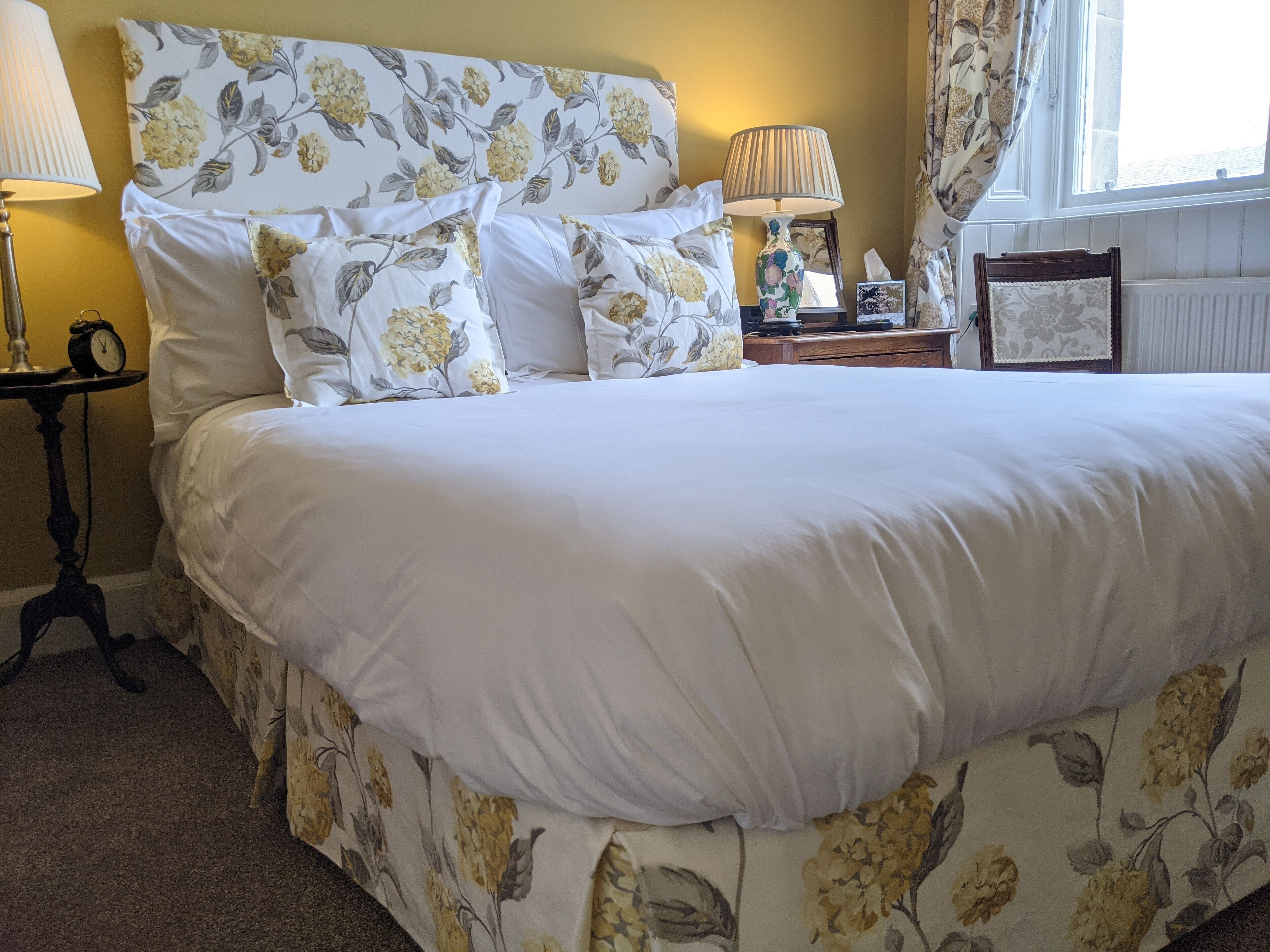 Una cama bien hecha con ropa de cama floral, una mesita de noche con una lámpara y una mesita con un reloj.