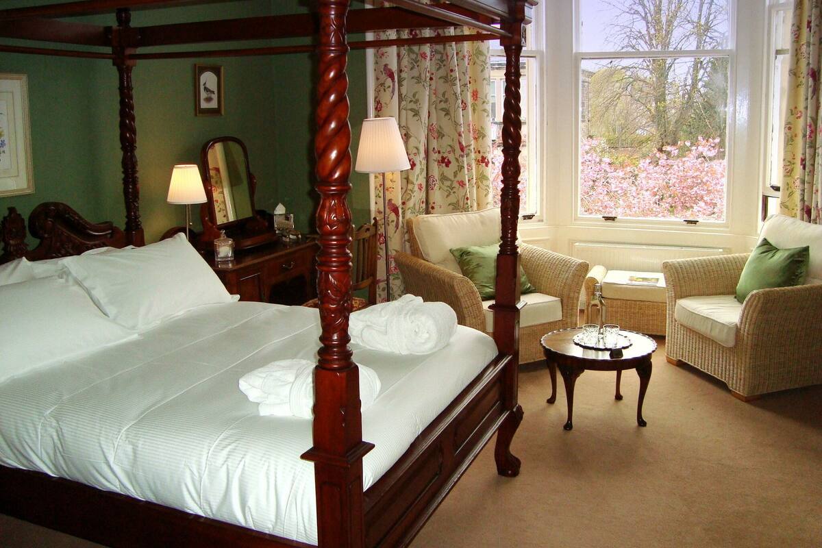 A four-poster bed with white linens, a side table with a lamp, and a view of blooming trees through a window.