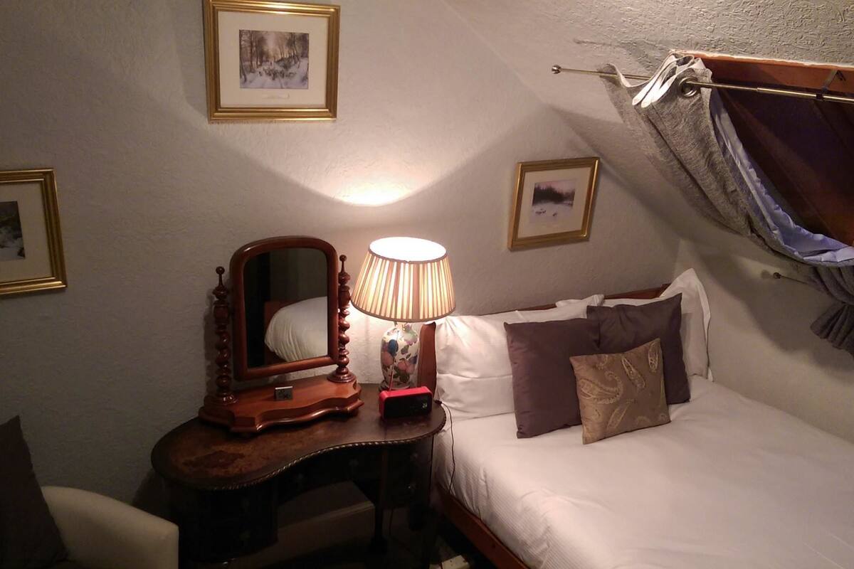 A bedroom with a wooden nightstand, a mirror, a lamp, a bed with white bedding and multiple pillows, and two framed pictures on the wall.