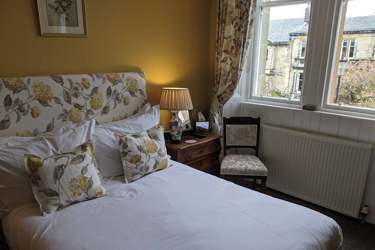 A neatly made bed with floral-patterned pillows, a bedside lamp, a chair, and a window with curtains.