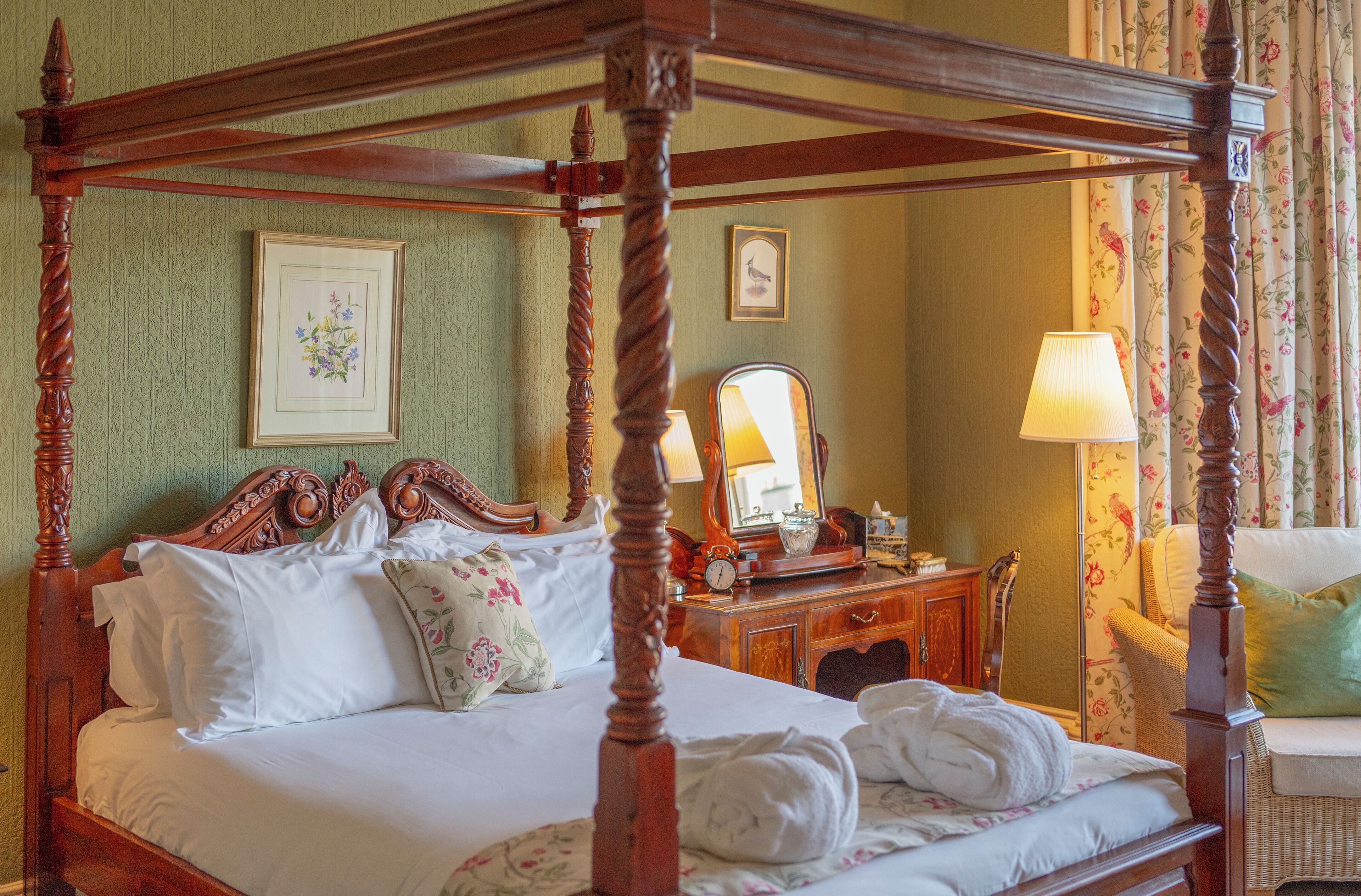 A four-poster bed with a wooden frame, a bedside table with a mirror, and a floral-patterned chair.