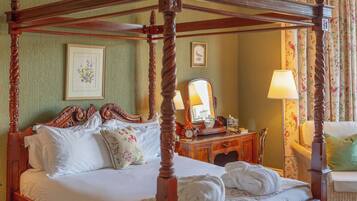 A four-poster bed with a wooden frame, a bedside table with a mirror, and a floral-patterned chair.