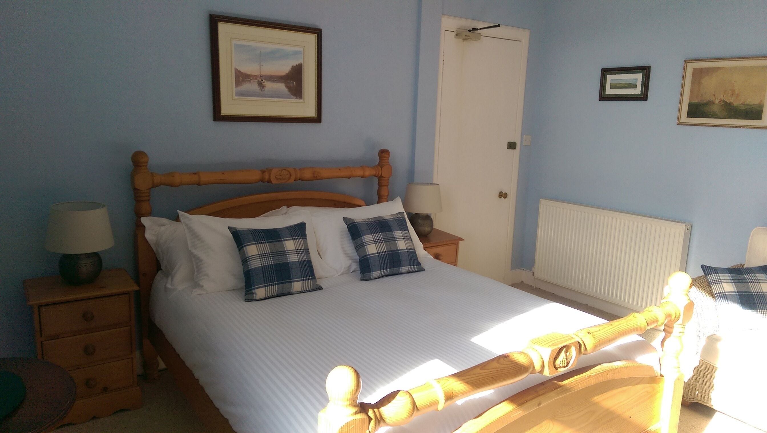 Une chambre à coucher avec un lit en bois, une literie blanche, deux coussins à carreaux, une table de chevet avec une lampe et un tableau encadré au mur.