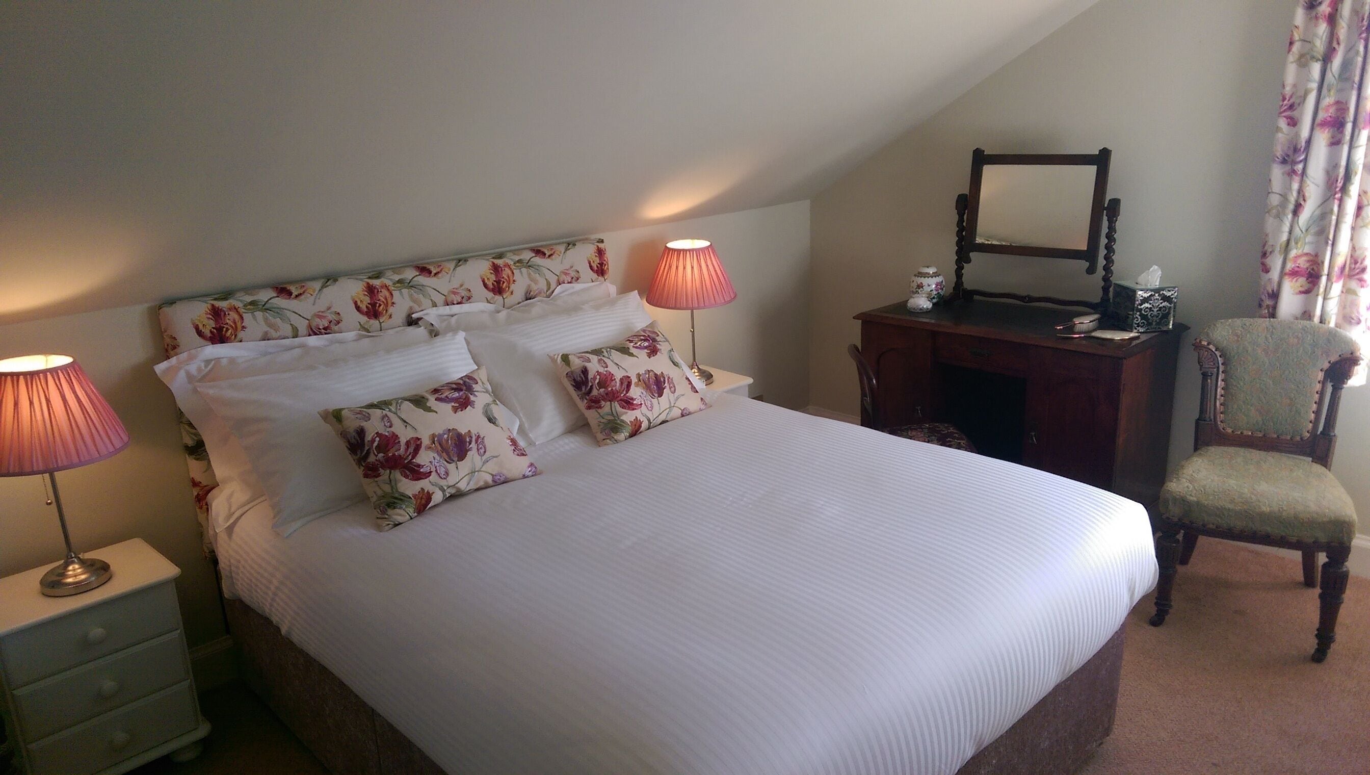 Cama bem arrumada com almofadas estampadas e duas luminárias de cabeceira. Apresenta uma mesa de cabeceira de madeira com um espelho e uma cadeira no canto.
