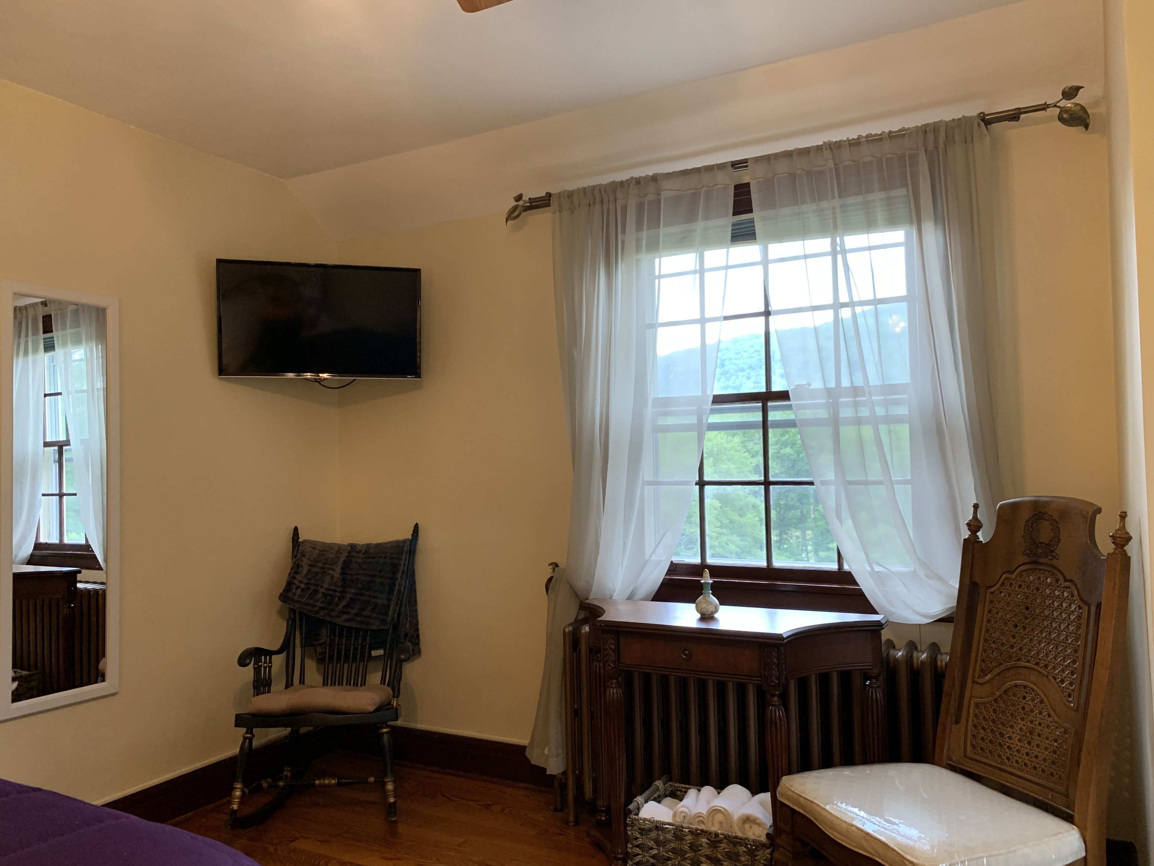 A room with a wooden rocking chair, a small table with a vase, a radiator, and a television mounted on the wall.