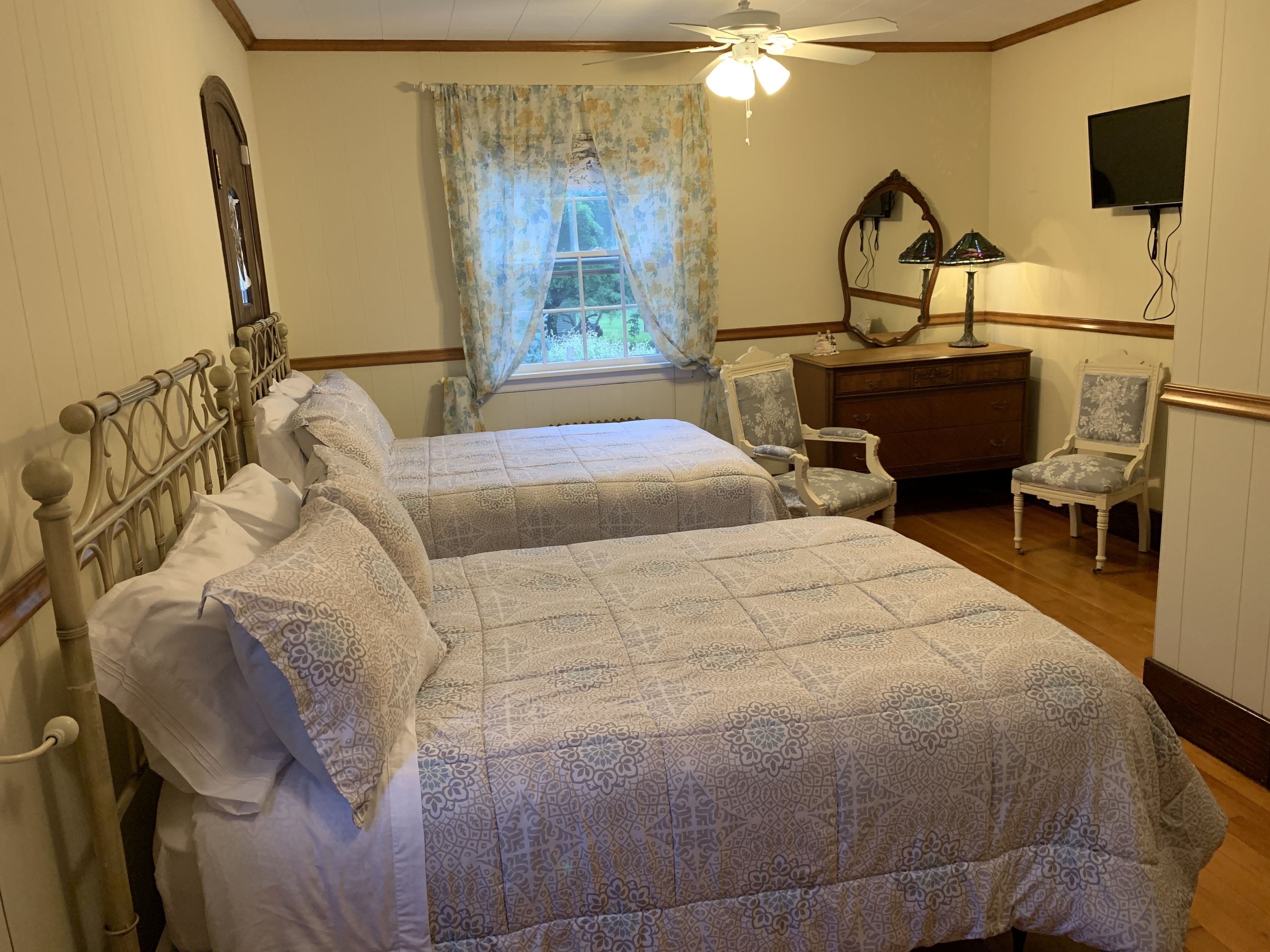 Une chambre avec deux lits, un ventilateur de plafond, une fenêtre avec des rideaux, une commode, une chaise et un téléviseur fixé au mur.