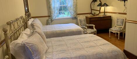 Une chambre avec deux lits, un ventilateur de plafond, une fenêtre avec des rideaux, une commode, une chaise et un téléviseur fixé au mur.