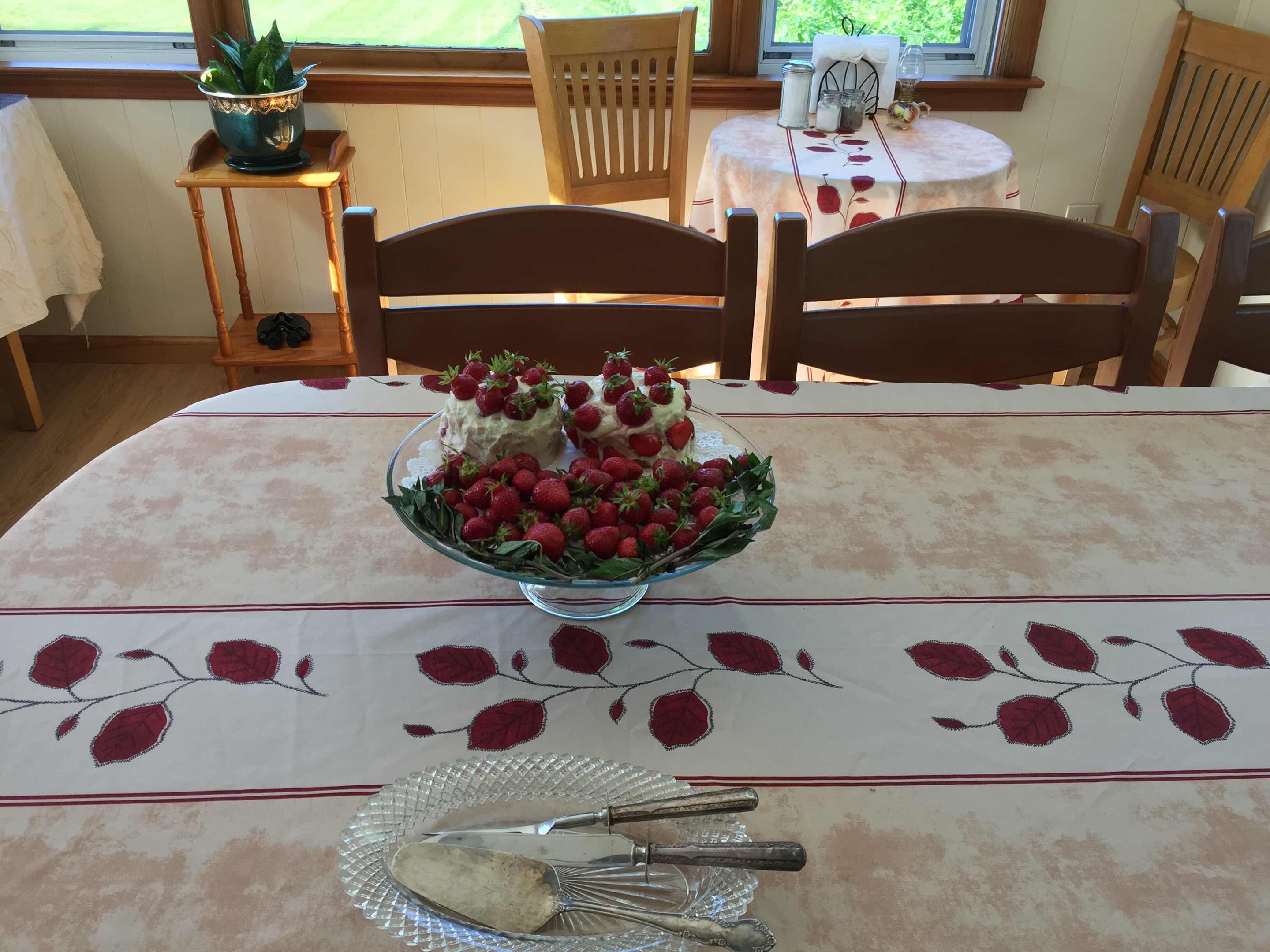 A dining table with a floral tablecloth, a glass bowl with strawberries, and a set of cutlery.