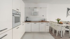A modern kitchen with white cabinetry, a built-in oven, a microwave, and a dining area with a wooden table and white chairs. - ShortStayPoland Aleje Jerozolimskie B13 (Warsaw)