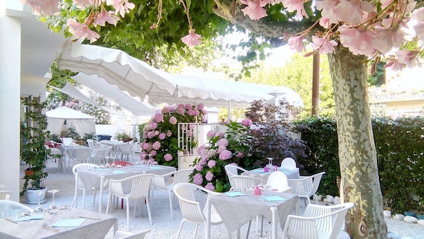 Un área de estar al aire libre con mesas y sillas blancas, rodeada de follaje y un arco decorativo de flores.